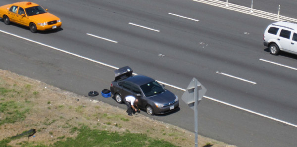 Still image from a traffic camera feed showing a man changing a flat tire of a vehicle parked on the shoulder of the highway.