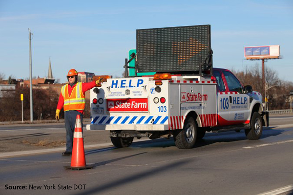 In the photo a man in a safety vest stands behind a New York State DOT HELP truck motioning motorists to merge right. The truck is installed with a large dynamic message sign, in this photo the sign shows a large arrow pointing to the right.