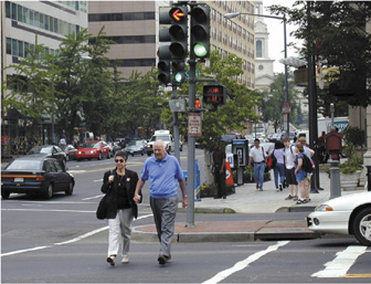 Photograph showing a man and woman holding hands as they cross a street at a busy, downtown intersection.