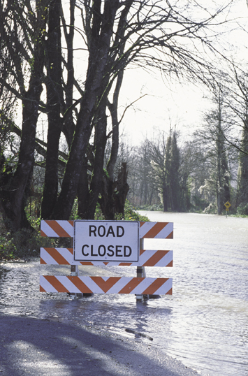 Photograph showing a road that has been flooded with water. A sign is posted in the roadway with