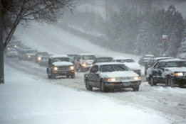 Photograph showing two lanes of vehicles traveling on a roadway in a snow storm. 
