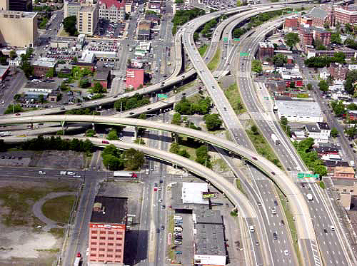 aerial photo of a freeway-to-freeway interchange in an urban setting, showing multiple ramps and infrastructure elevations. One freeway is shown aligned vertically and the other horizontally, with two levels of flyovers above urban streets