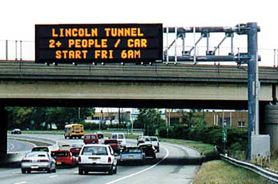 photo of a freeway approaching the entrance to the Lincoln Tunnel. A changeable message sign states: "LINCOLN TUNNEL, 2+ PEOPLE / CAR, START FRI 6AM"