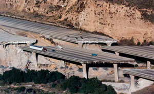 This is an aerial picture of a freeway bridge damaged in the Northridge earthquake in January 1994 that was completely torn away from the main part of the highway. The bridge has been separated from the freeway on both sides of the bridge, and a tractor-trailer, bus, and cars are still visible on the bridge. 