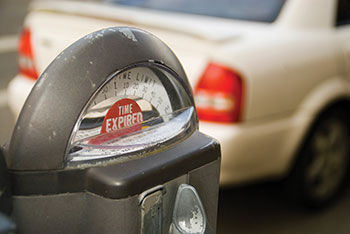Photo. Close up of a parking meter showing “Time Expired,” with the rear of a car in the background.