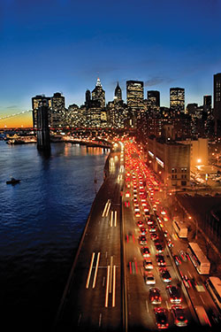 Photo. Nighttime view of the West Side Highway and downtown Manhattan, including the Hudson River. Image shows a steady flow of traffic along the highway.