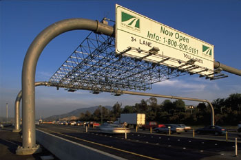 Photo. Transponders are read by overhead antennas, allowing tolls to be paid without stopping. Highway traffic passes under the overhead antennas and a sign that indicates a split in highway lanes (“3+ lanes” and “toll lanes”).