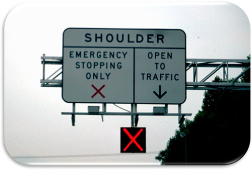 Figure 16. Photo. I-66 Regulator Sign with Lane Control Signals (Photo Courtesy of VDOT). Photo showing an overhead regulatory sign with a lane control signal used in Virginia to indicate active temporary shoulder use. A banner over the sign reads "SHOULDER". The left half of the sign reads "EMERGENCY STOPPING ONLY" with a red X. The right half of the sign reads "OPEN TO TRAFFIC" with a down arrow. The lane control signal shows a red X indication.
