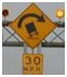 Photograph. A yellow diamond-shaped road sign depicting a sharp curve with a truck possibly overturning and cautioning 30 miles per hour represents “changes in highway alignment.”