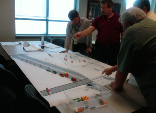 A photo of NYSDOT TIM Responder Training with a small group of people standing at a table and looking at a response situation presented on a small-scale map of a roadway with miniature vehicles.