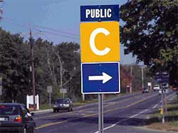 photo showing a guide sign, mounted on a post located on the right shoulder of a three-lane roadway, stating "public" followed below by a large "C" and a right arrow