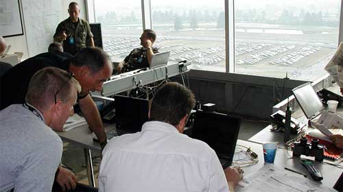 photo of several men bending over computer monitors and display consoles in an office with large windows overlooking a parking lot filled with cars