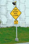Photograph - Advance warning sign with warning lamp, posted on left hand side of roadway.  The sign reads RAMP METERED WHEN FLASHING and is positioned immediately below the warning lamp and above another smaller sign that reads FORM TWO LINES.  Both signs are yellow, the larger of the two is a diamond and the smaller of the two is a rectangle.