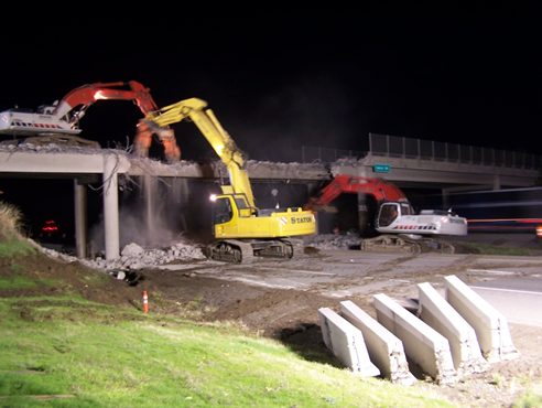 Image shows three heavy backhoes demolishing an overpass at night.