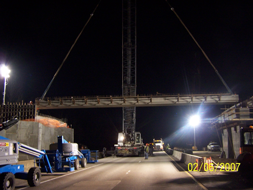 Image shows a crane lowering a pre-fabricated bridge section into place at night.
