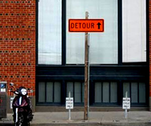 Photo of a Detour warning sign with an upward pointing arrow mounted at the top of a pole on a sidewalk.