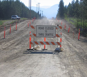 Photo of a white sign displaying Road Closed to Thru Traffic mounted on a three-rail barricade and blocking an unpaved roadway lined on both sides with tubular markers.