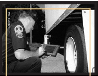 Photograph of a CVISN inspector seated near a truck's rear wheel during safety inspection and using wireless, handheld technology.