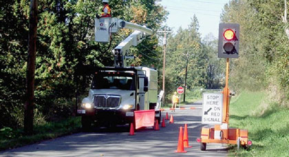 This photo shows an automated flagger assistance device set up on the right side of a small roadway, while a maintenance truck and crew works on powerlines on the left side of the roadway.
