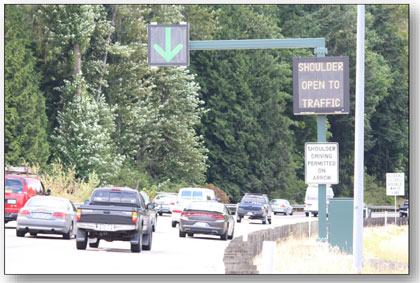 Photograph of the dynamic signing used in Seattle to indicate the shoulder is open to traffic.  A DLUC is illuminated with a green downward arrow over the shoulder and a small DMS on the shoulder reads 'SHOULDER OPEN TO TRAFFIC.'  A statiC regulatory sign reads 'SHOULDER DRIVING PERMITTED ON ARROW.'