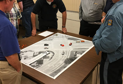 People looking at a map of a multi-lane four-way intersection. On top of the map miniature cars, an ambulance and other emergency service vehicles are placed showing a scenario for a traffic incident.