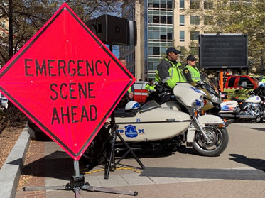 A large diamond-shaped sign sitting on the sidewalk that says Emergency Scene Ahead and two police officers stand behind it next to their motorcycles.