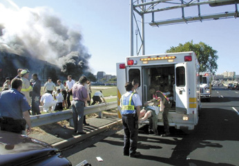 Photograph from September 11, 2001, showing the exterior of The Pentagon, with smoke coming from the building and a person being loaded into an ambulance while several people look on.