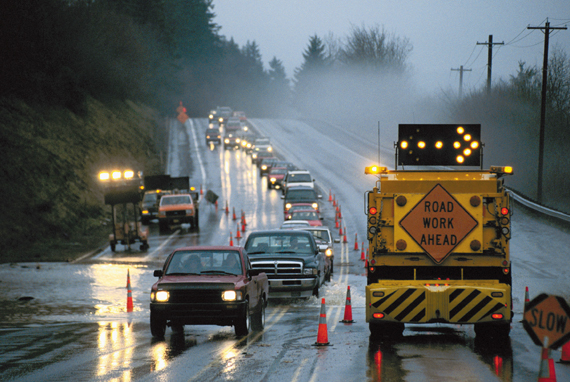 Photograph showing vehicles traveling through a work zone in one lane. The other lane is blocked by a flashing arrow signal directing traffic to move to the right and a sign with the words, "Road Work Ahead" on it. 