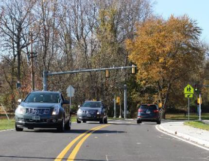 An image with three cars – two in one lane, and one approaching a crosswalk in the other on a fall day.