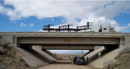 An image of bridge under construction.  Underneath the bridge are workmen and cars, while on the bridge is a truck and work equipment.