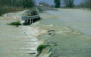 Figure 3-8 Flooding Damages ITS as well as Structures: water from an overflowing river washes over a two-lane road. The road edges have started to decay and soil is being eroded from the area