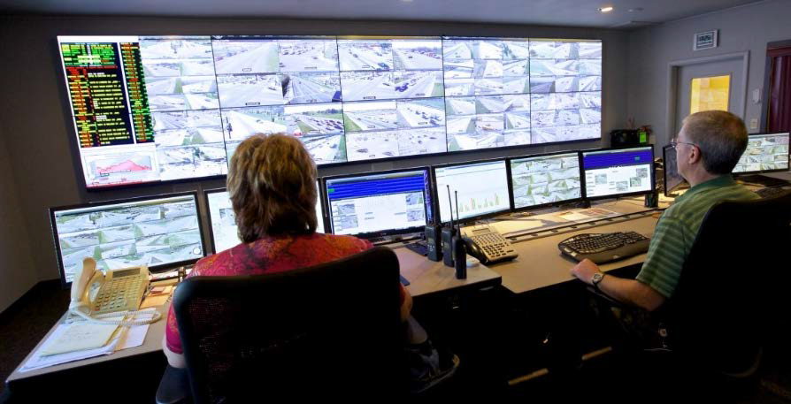 Image of ACHD Transportation Management Center with two persons sitting at a desk with numerous screens of traffic being observed.