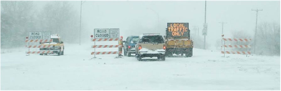 Snowy Road with road closed barricades.  A sign mounted on the back of a work truck reads 'Local Traffic Only'.