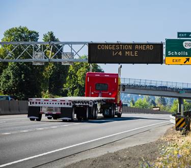 Image of a red flatbed freight truck on an interstate.  Overhead is a sign that says there is congestion ahead in one-fourth of a mile.