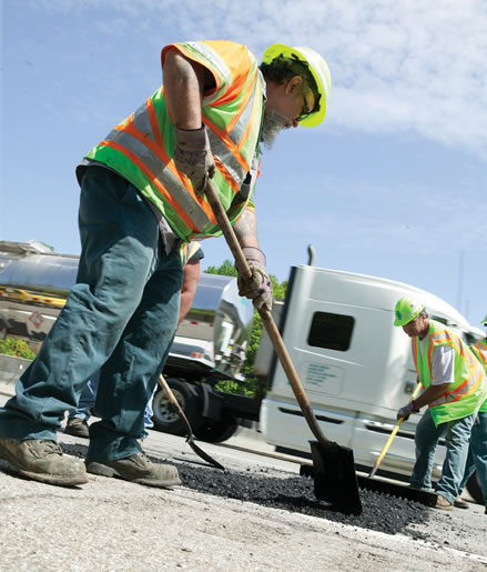 Image is of workmen in orange and yellow vests with shovels filling potholes on a road.