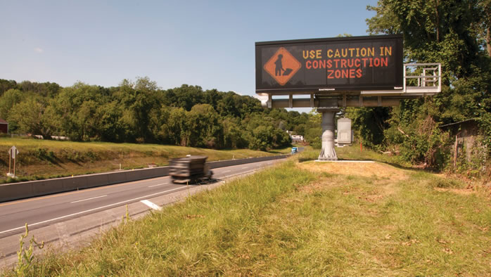 The image is of a highway with median with a truck on the road and a large electronic road sign that reads: Use Caution in Construction Zones.