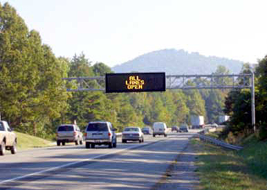 photo of overhead light-emitting matrix message sign mounted over the center of one side of a divided highway with the message "ALL LANES OPEN"