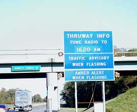photo of a static HAR sign with flashing beacons installed on both sides of a sign with a green background and white lettering. The sign is shown outside a highway shoulder and shows the message "THRUWAY INFO, TUNE RADIO TO 1620 AM, TRAFFIC ADVISORY WHEN FLASHING, AMBER ALERT WHEN FLASHING"