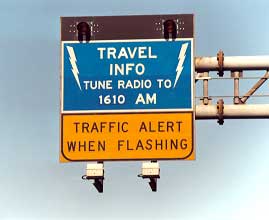 photo of a static HAR sign with flashing beacons on top of a sign with a green background and white lettering. The sign is shown as an overhead sign and shows the message "TRAVEL INFO, TUNE RADIO TO 1610 AM". A yellow advisory plaque mounted below the sign shows the message "TRAFFIC ALERT WHEN FLASHING"