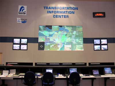 photo of a video display wall in a TMC, showing a large map panel and two four-screen video panels; a large multi-station console and chairs are shown facing the display wall