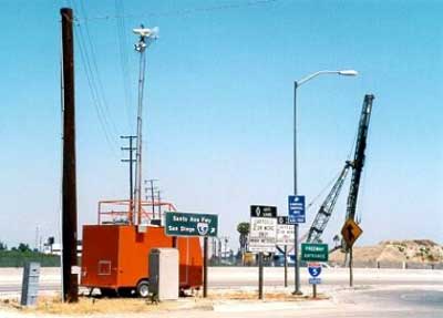 photo of a CCTV mounted at the top of a tall telescoping boom attached to a trailer; it is shown grouped with a telephone pole, four sign assemblies, an overhead street light, and two cranes next to a roadway