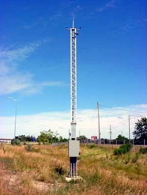 photo of an environmental sensor station, showing vertical and horizontal instruments mounted at the top of a tall narrow frame on top of a base cabinet; it is shown in a field near a roadway