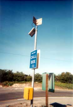 photo of a call box installation, showing a box that contains a telephone mounted on a pole adjacent to a roadway, with a sign ("CALL BOX" and location) mounted above it and three solar panels mounted at the top of the pole