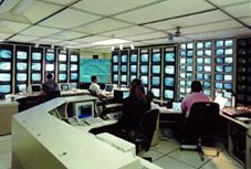 Photo of traffic management center with workers at work stations surrounded by walls of video monitors.