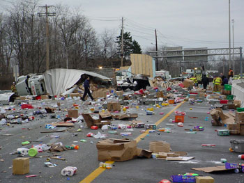 A picture of a roadway accident. A tractor trailer has been ripped in half and overturned in the background. The foreground shows the roadway littered with the contents of the truck (toilet paper rolls, detergent bottles, and assorted other boxes and packages). All lanes of the roadway are obstructed, both by the wreck of the truck and its contents. In the far background, emergency response personnel are surveying the scene, but cleanup has not yet begun.