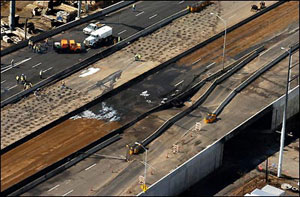 A photograph of the I-95 bridge collapse taken from the air. At least three lanes (one half of the bridge capacity) and one exit ramp are visibly sagging, and the pavement appears scorched and blackened. The bridge is not currently open to traffic, as the few lanes that appear intact are occupied by what appear to be construction personnel and a handful of vehicles.