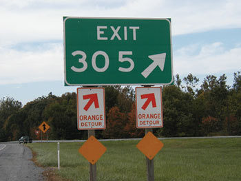 A group of road signs. The top sign is a green rectangle with white lettering, reading “Exit 305,” with an arrow pointing to the upper right. Below that, parallel to each other at either corner, are two white rectangular signs with orange lettering, each reading “Orange detour,” with arrows above the text pointing to the upper right. Below each of those signs is an orange diamond sign with no lettering. The background depicts a roadway with trees, identifiable as a merge lane by a yellow diamond arrow sign and an inverted triangular “Yield” sign.