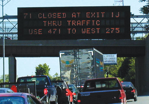 Photograph of highway traffic, showing traffic moving underneath an overpass with a programmable marquee signboard for traffic advisories. The text for the signboard reads “71 closed at exit IJ, thru traffic use 471 to west 275.” 