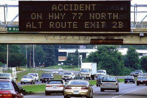 Photograph of highway traffic, showing traffic moving underneath an overpass with a programmable marquee signboard for traffic advisories. The signboard reads &ldquo;Accident on hwy 77 north, alt route exit 2B.&rdquo;