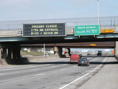 A photograph of highway traffic flow underneath an overpass. Mounted on the overpass, next to two standard highway signs, is a programmable marquee traffic advisory signboard reading “Freeway closed, I-96 WB express, M-39/Outer Dr.”